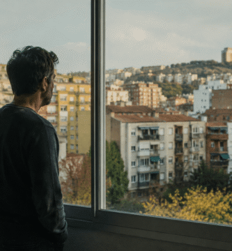 Hombre observando una zona residencial desde una ventana mientras reflexiona sobre una inversión inmobiliaria.